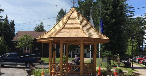A wooden gazebo under construction, surrounded by trees and flags, with workers and a truck nearby.