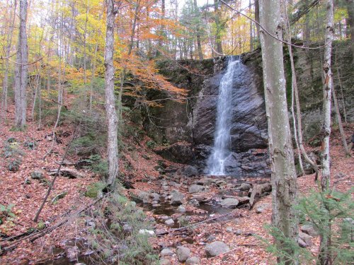 A serene waterfall cascades down rocky cliffs, surrounded by autumn foliage and a forest of trees.