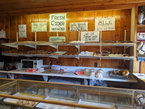 A cozy bakery display with shelves of baked goods, signs for cider, iced coffee, and various types of bread and donuts.