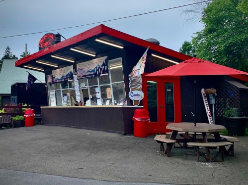 A small ice cream stand with a red roof, outdoor seating, and colorful signage in a wooded area.