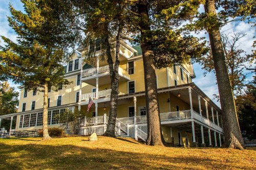 A large yellow building with multiple stories, surrounded by trees and a grassy area, featuring a porch and an American flag.