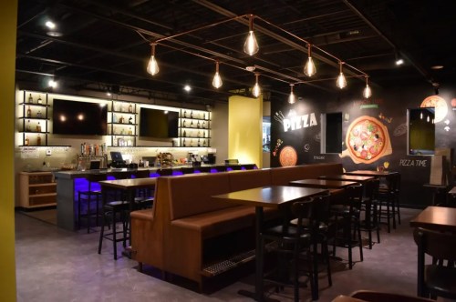 Modern pizza restaurant interior with a bar, wooden seating, and stylish lighting. Walls feature pizza-themed decor.