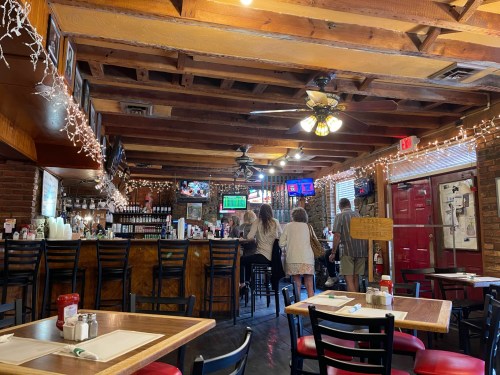 A cozy restaurant interior with wooden beams, a bar, and patrons seated at the counter, decorated with string lights.
