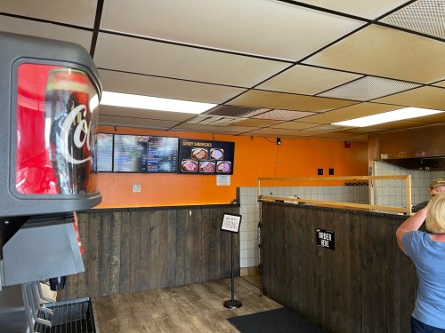 Interior of a casual eatery with a soda machine, menu board, and a sign indicating where to order. Warm orange walls.