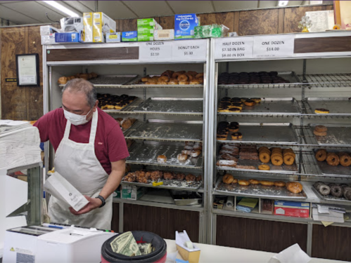 A masked baker prepares donuts in a shop with shelves displaying various baked goods.