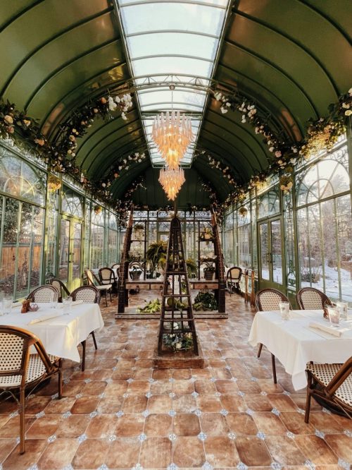 A beautifully decorated greenhouse with tables, chairs, and a chandelier, surrounded by plants and floral accents.