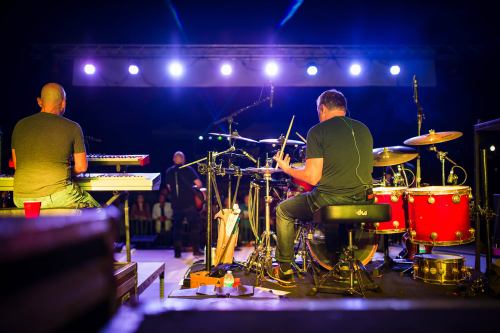 Two musicians perform on stage, one at a keyboard and the other on drums, with colorful lights illuminating the scene.