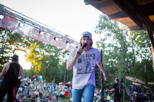 A singer performs on stage at an outdoor concert, with a crowd and trees in the background.