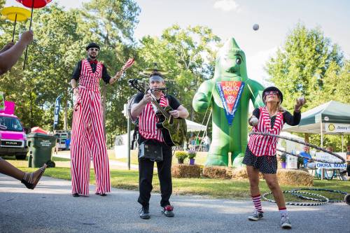 A lively festival scene with performers in striped outfits, one on stilts, playing guitar, and another hula hooping near a large inflatable.