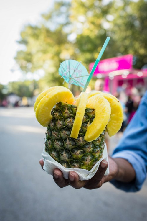 A person holds a pineapple cup filled with a drink, garnished with pineapple slices and a small umbrella.