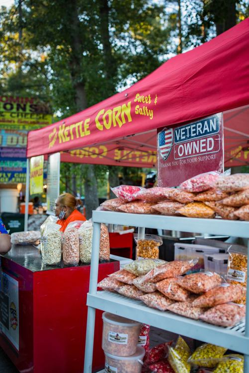 A vendor booth with a red canopy selling kettle corn, featuring bags of popcorn and a "Veteran Owned" sign.
