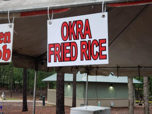 Sign reading "Okra Fried Rice" hanging at a food stall in a wooded area.