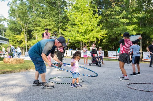 A man helps a young child hula hoop at a lively outdoor event with performers and families in the background.