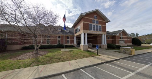 Exterior view of a brick library building with a flagpole, surrounded by greenery and a parking lot.