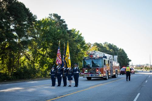 A fire truck and emergency vehicles parade past a group of uniformed officers holding flags on a tree-lined street.