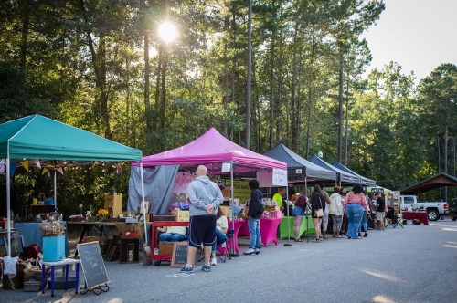 A vibrant outdoor market with colorful tents, vendors, and shoppers amidst tall trees and sunlight.