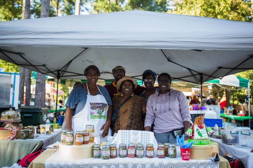 A group of five people smiles behind a market stall filled with jars, under a large tent at an outdoor event.