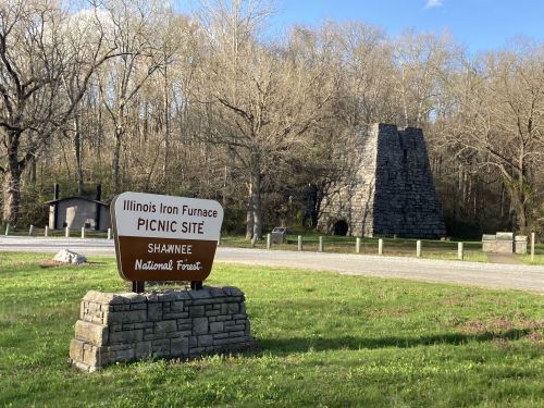 Sign for Illinois Iron Furnace picnic site in Shawnee National Forest, with a stone furnace structure in the background.