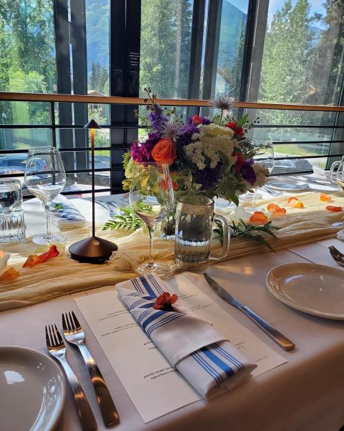 A beautifully set dining table with flowers, glassware, and a menu, overlooking a scenic view through large windows.