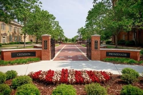 A brick pathway named "Crimson Promenade" lined with greenery and flowers, leading through a campus area.