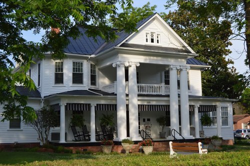 A large white house with a porch, columns, and rocking chairs, surrounded by greenery and flower pots.