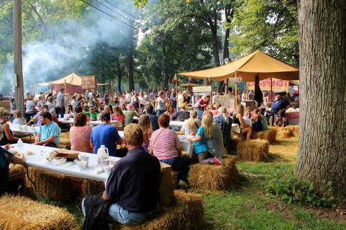 A lively outdoor gathering with people dining at tables surrounded by hay bales, food stalls, and trees.
