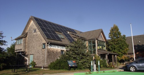 A wooden building with solar panels on the roof, surrounded by trees and a clear blue sky.