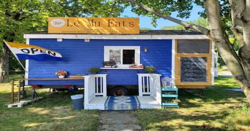 A blue food truck with a yellow sign reading "Le Mu Eats," featuring an open flag and outdoor seating.