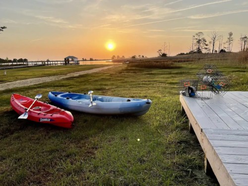 Two kayaks on grass by a dock at sunset, with a serene water view and a hint of trees in the background.