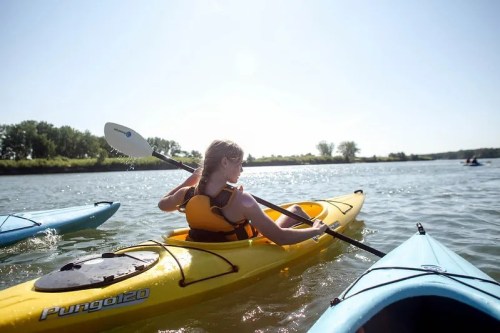 A person paddles a yellow kayak on a calm river, with other kayakers visible in the background under a clear sky.