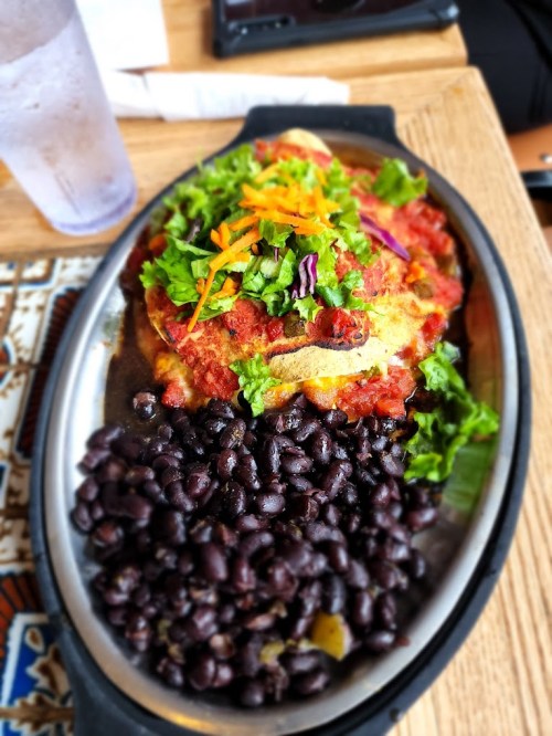 A sizzling plate of enchiladas topped with sauce, lettuce, and carrots, served with a side of black beans.