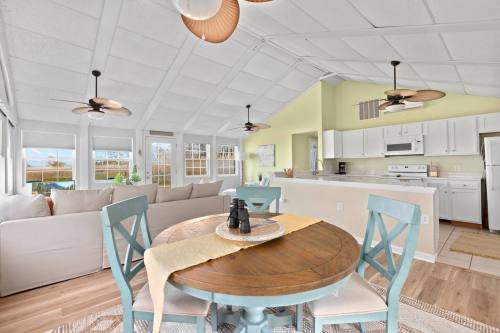 Bright, airy dining area with a round wooden table, blue chairs, and a cozy living space featuring large windows.