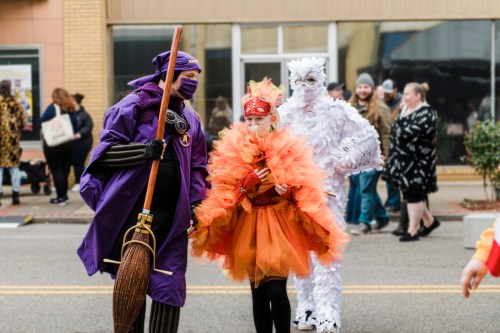 A colorful parade scene featuring a person in a purple costume with a broom, a child in an orange outfit, and a white figure.