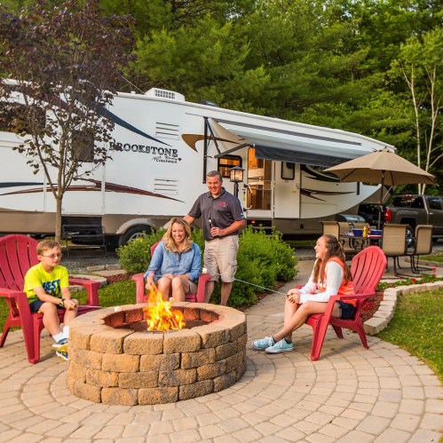 A family enjoys a campfire outside their RV, sitting in red chairs with trees in the background.