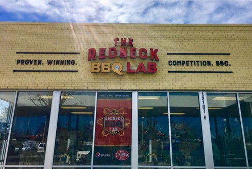 Sign for "The Redneck BBQ Lab" featuring a yellow brick exterior and large windows. Slogan: "Proven. Winning. Competition. BBQ."
