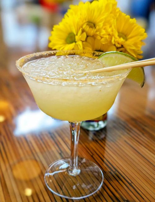 A refreshing margarita in a glass with a salted rim, garnished with a lime slice, next to a bouquet of yellow flowers.