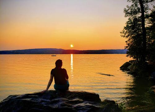 A person sits on a rock by the water, watching a sunset that reflects on the calm lake. Trees frame the scene.