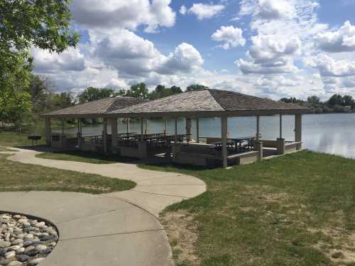 A lakeside pavilion with picnic tables, surrounded by grass and a walking path, under a cloudy sky.