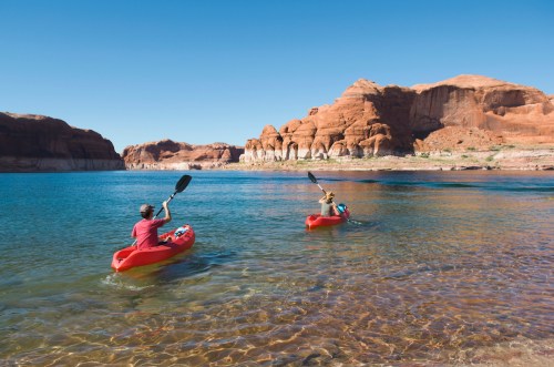 Two people kayak on a clear blue lake surrounded by red rock formations under a bright blue sky.