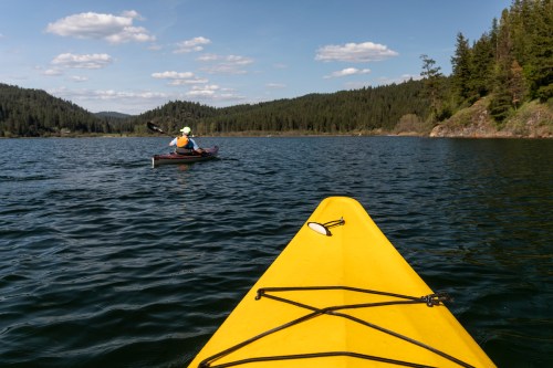 A yellow kayak in the foreground with a person paddling in a kayak on a calm lake surrounded by trees and hills.