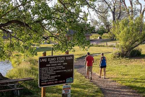 Two people walk along a path in Lake Elmo State Park, with a sign detailing park regulations nearby.