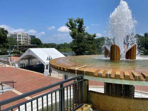 A fountain with water spraying, overlooking a park area with a tent and trees by a river under a blue sky.