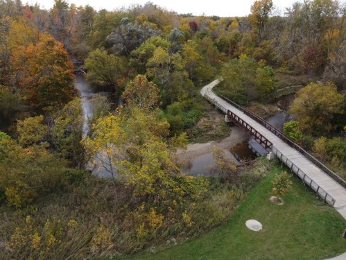 Aerial view of a winding river surrounded by trees in autumn colors, with a wooden bridge crossing over the water.