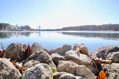 A serene lakeside view with smooth rocks in the foreground and trees lining the opposite shore under a clear blue sky.