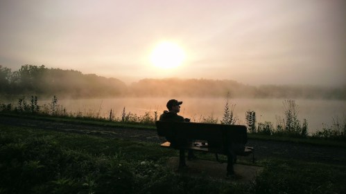 A person sits on a bench by a misty lake at sunrise, surrounded by trees and soft fog.