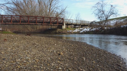 A peaceful river scene with a gravel bank, a wooden bridge in the background, and bare trees along the shore.