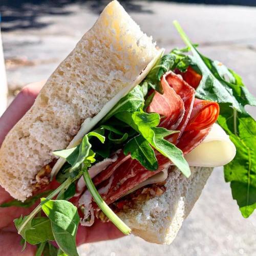 A hand holding a sandwich filled with leafy greens, sliced meats, and cheese, with a soft white bread exterior.