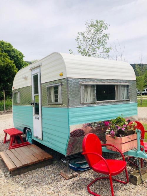 A vintage teal and white camper trailer with red chairs and flowers, set in a green outdoor area.