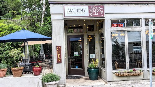 Exterior of Alchemy Bistro & Wine Bar, featuring an "OPEN" sign, outdoor seating, and potted plants.