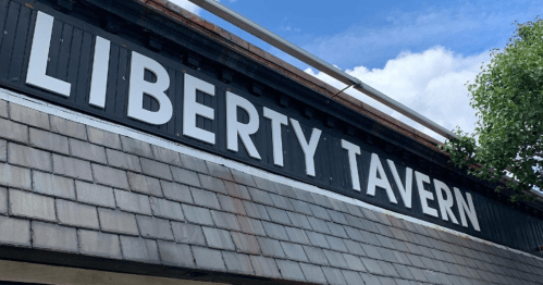 Sign for Liberty Tavern against a blue sky with clouds, featuring a wooden shingle roof.